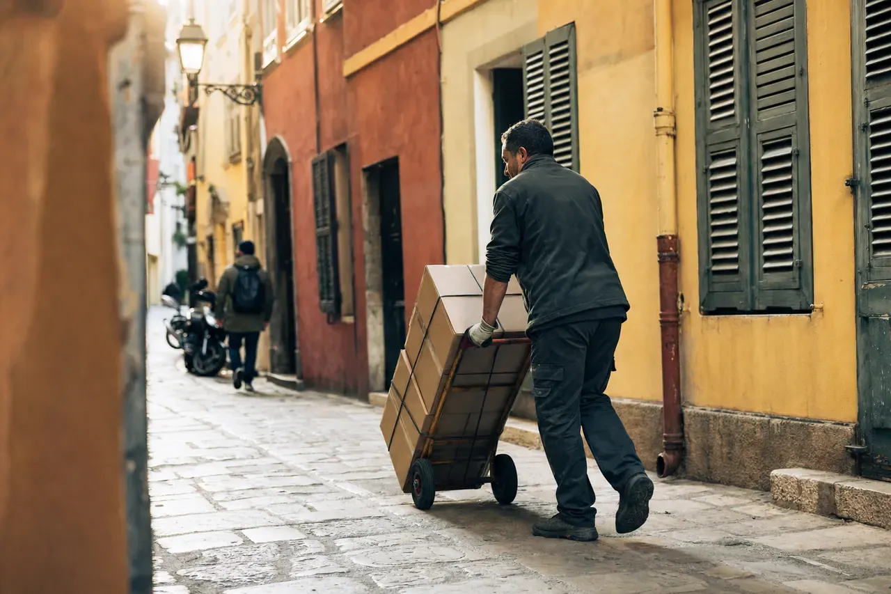 Un diable chargé de cartons sanglés poussé dans une ruelle du Vieux-Nice aux façades colorées